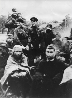 Grupa jeńców sowieckich grzejących się przy zapalonym ognisku. Niemiecki wartownik uzbrojony w karabin w płaszczu i furażerce.